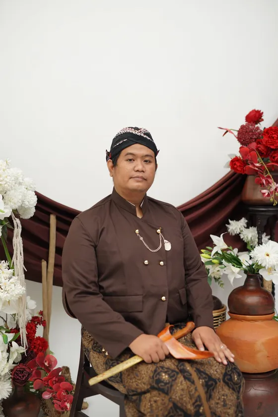 Mempelai pria dalam beskap cokelat dan blangkon, duduk memegang keris