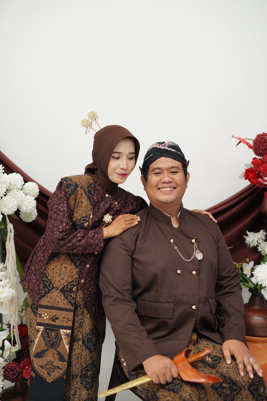 Satrio dan Maryam dalam busana adat Jawa berwarna cokelat, Maryam berdiri di belakang Satrio yang duduk